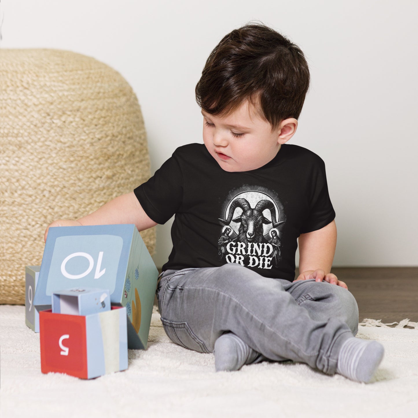 A toddler in a Bougie Boiz Club Grind Or Die baby t-shirt, featuring a white rams head graphic, stands against a plain white background. With short blonde hair and blue jeans, this determined little lifter already exudes style.