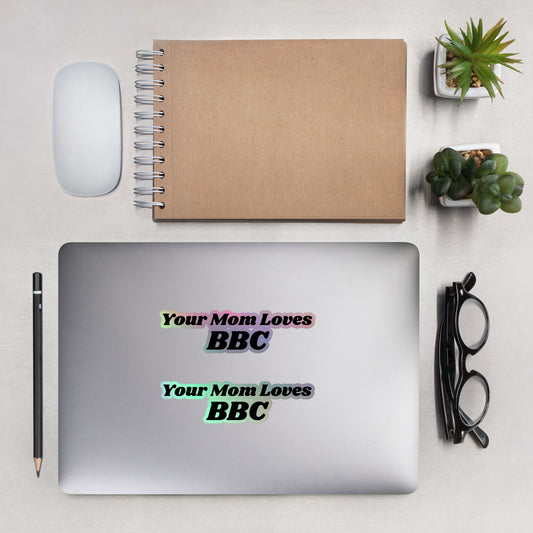 A neatly arranged desk features a silver laptop decorated with Bougie Boiz Clubs Your Mom Loves BBC holographic stickers. Nearby, you’ll find a spiral-bound notebook, mouse, pencil, eyeglasses, small potted plants, and a piece of paper.