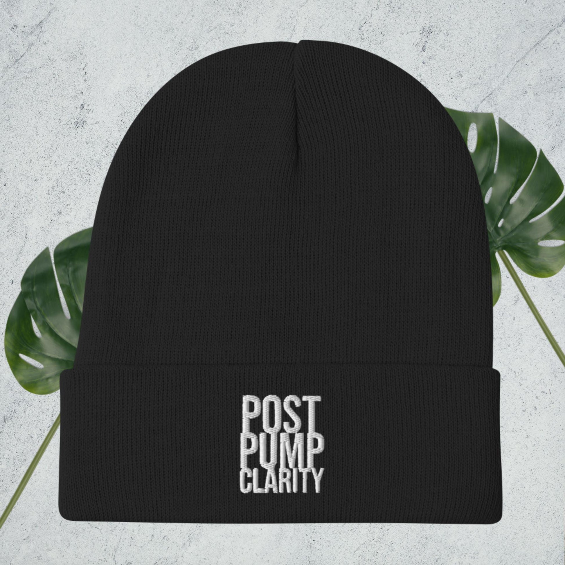 The Bougie Boiz Club Post Pump Clarity black beanie, ideal for the gym, features white embroidery on the front and rests against a textured surface with lush green monstera leaves partially in view.