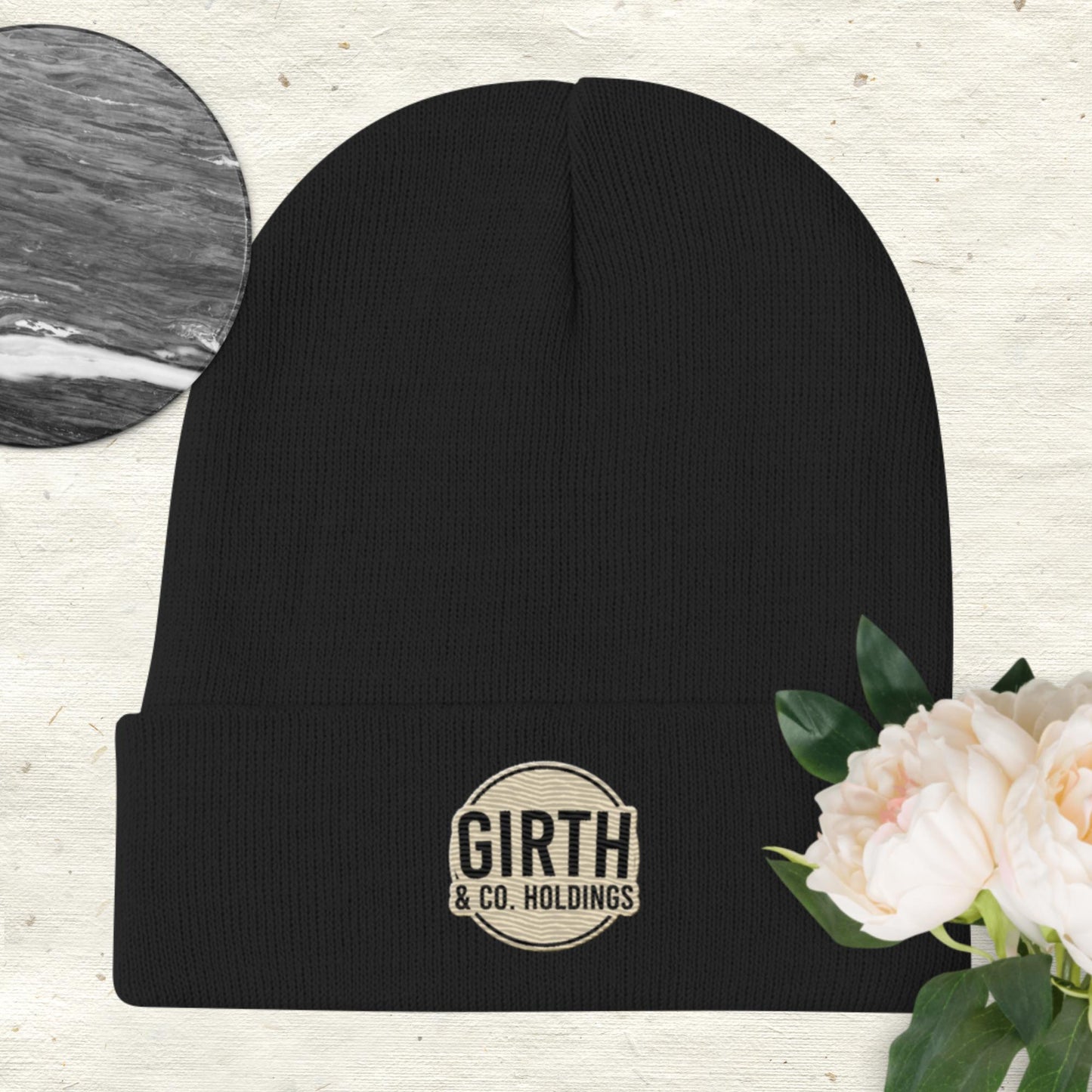 A cozy Bougie Boiz Club green beanie, embroidered with the Girth & Co. logo, sits on a textured backdrop of black and white floral elements, accented by pink and white peonies blooming in the lower right corner.