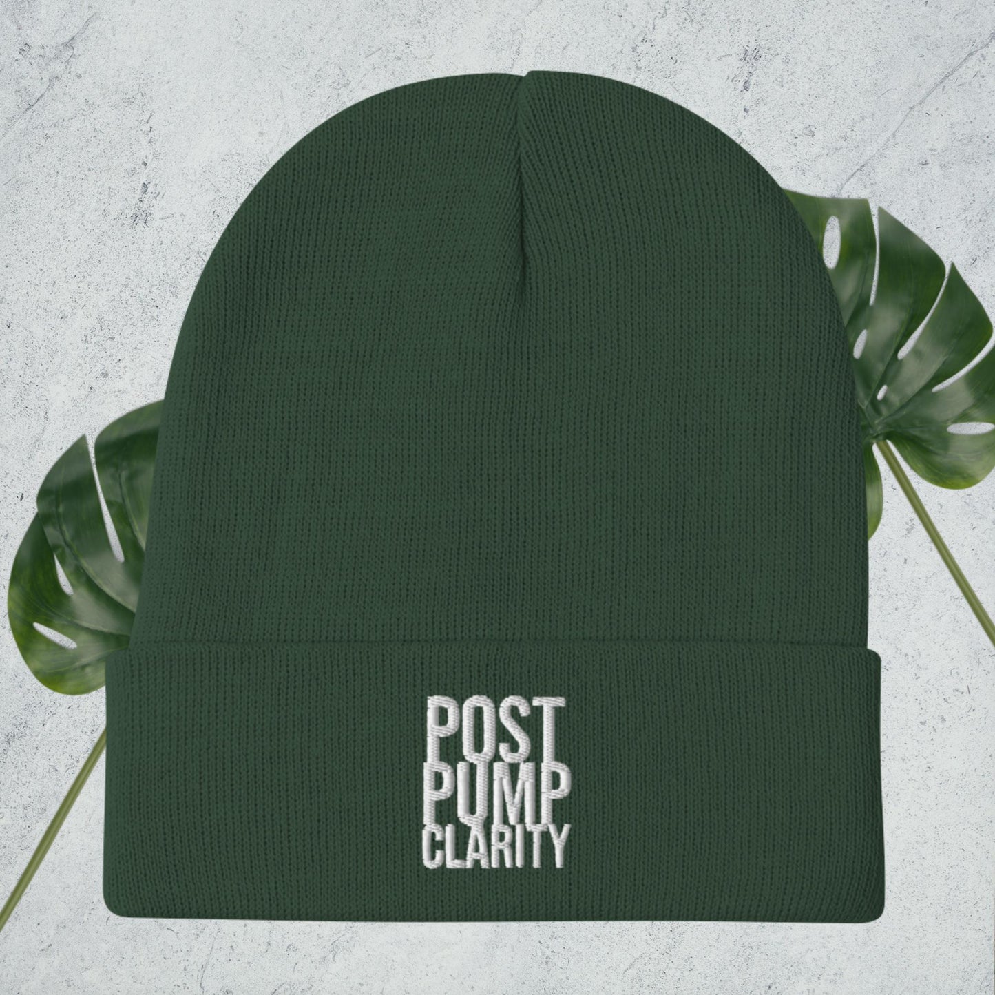 The Bougie Boiz Club Post Pump Clarity black beanie, ideal for the gym, features white embroidery on the front and rests against a textured surface with lush green monstera leaves partially in view.