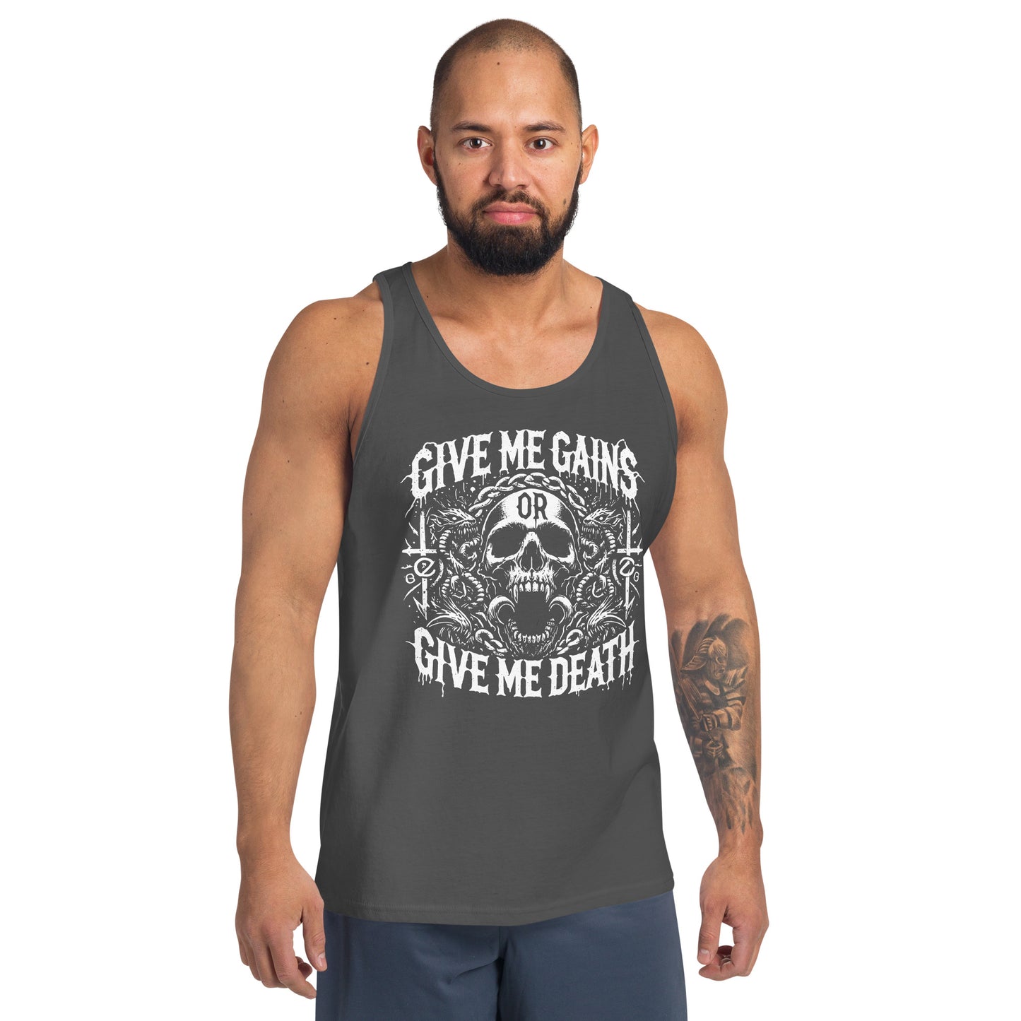 A bearded man embraces the iron life in a Bougie Boiz Club Give Me Gains - Tank Top featuring a skull graphic. Against a plain white backdrop, he pairs this black top with blue shorts, exuding serious gym vibes.