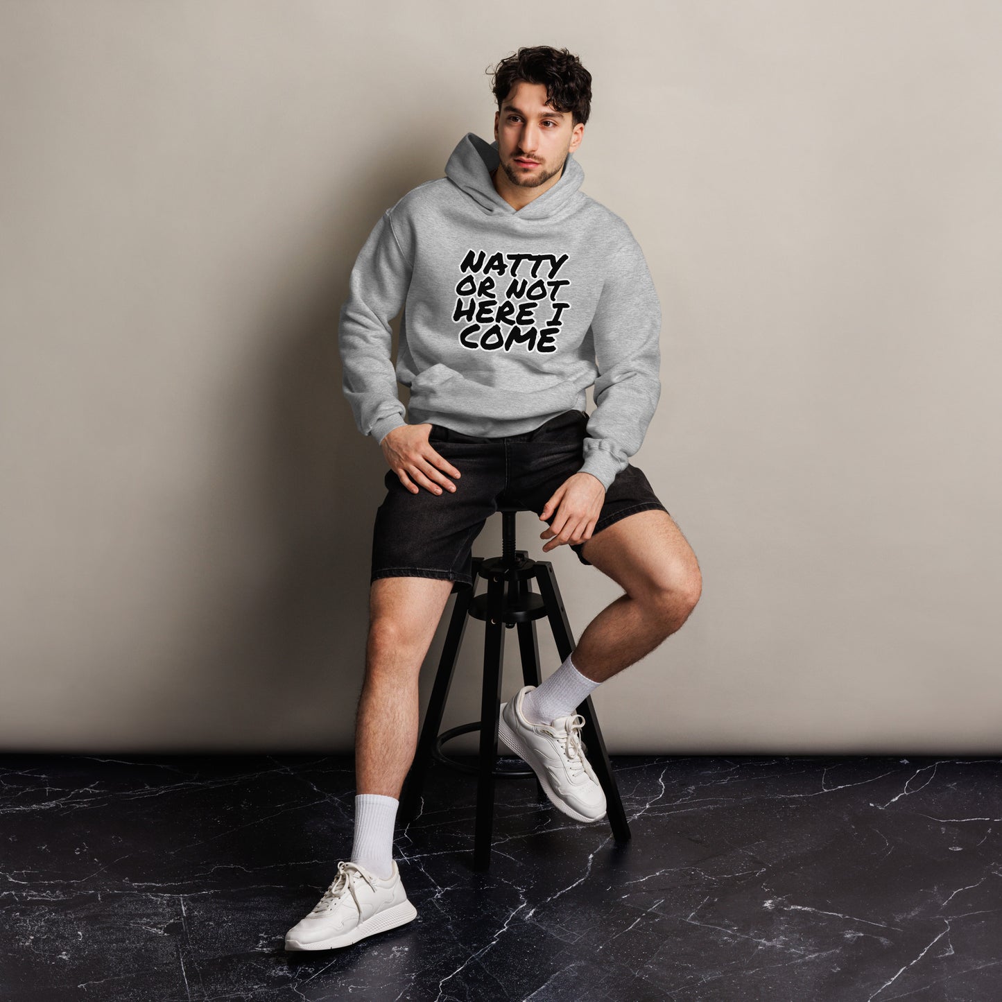 A man sits on a stool against a plain wall, wearing a Bougie Boiz Club Natty Or Not - Unisex Oversized Hoodie with the text READY OR NOT, HERE I COME. The fit complements his black shorts, white socks, and sneakers. The floor beneath displays a subtle marble pattern.