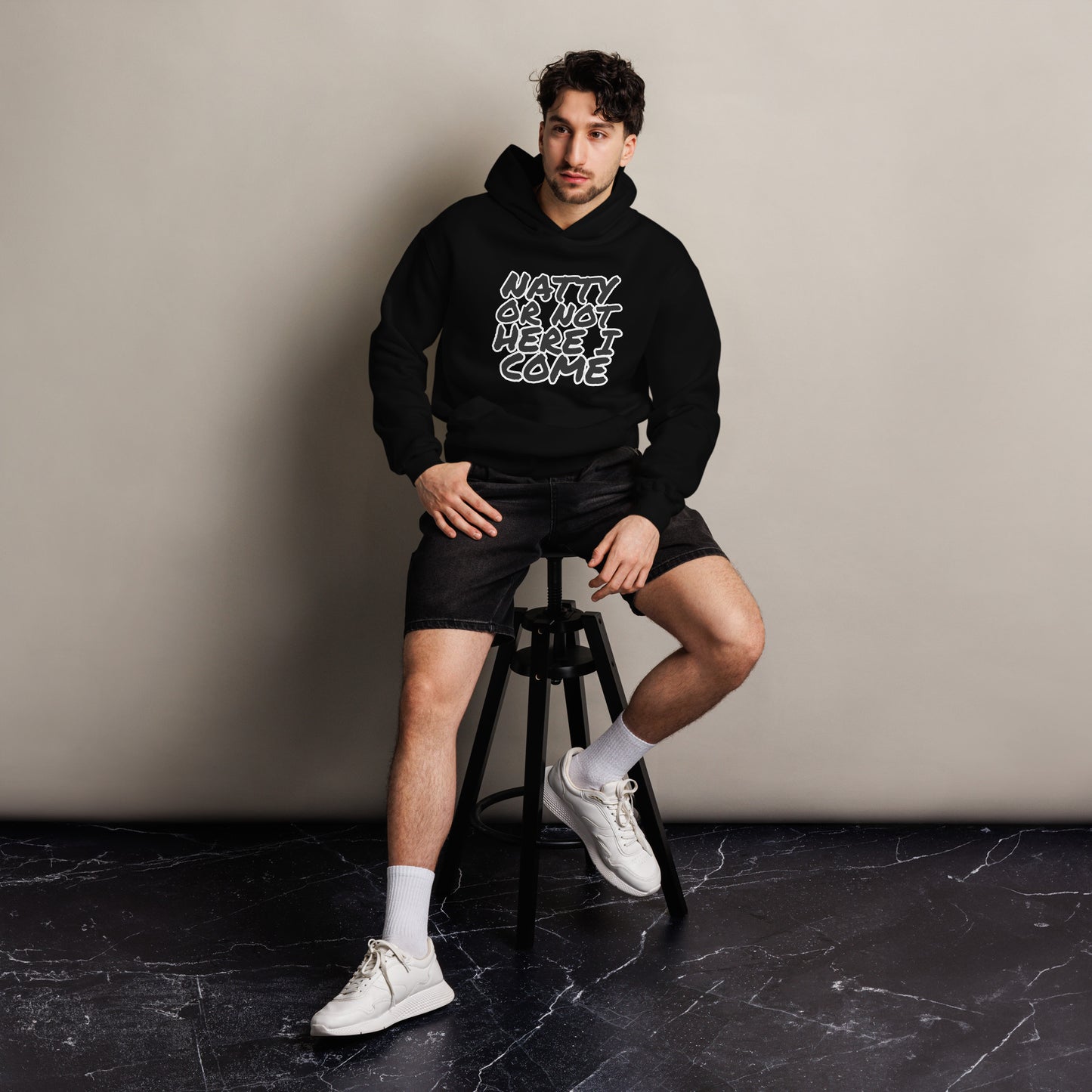 A man sits on a stool against a plain wall, wearing a Bougie Boiz Club Natty Or Not - Unisex Oversized Hoodie with the text READY OR NOT, HERE I COME. The fit complements his black shorts, white socks, and sneakers. The floor beneath displays a subtle marble pattern.