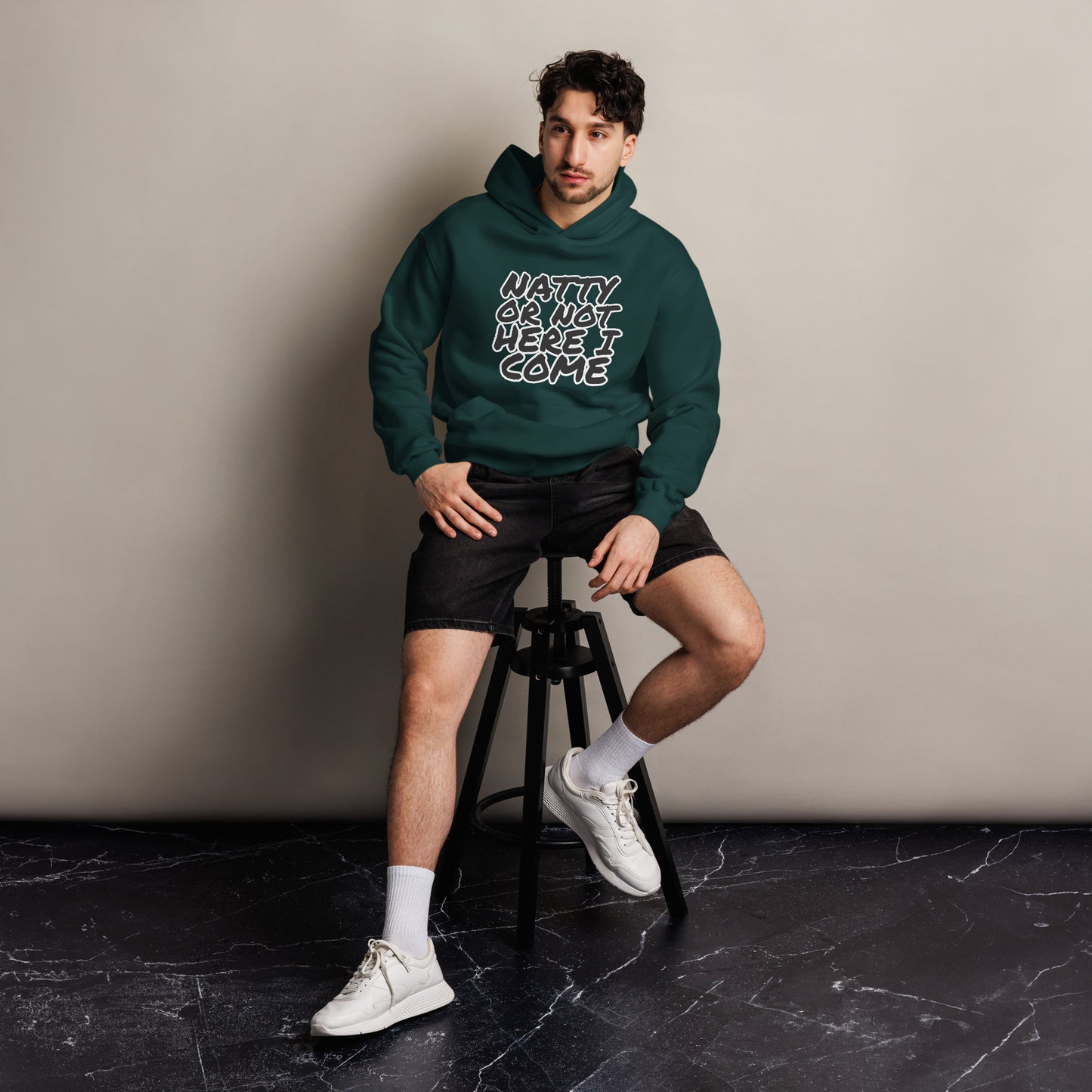 A man sits on a stool against a plain wall, wearing a Bougie Boiz Club Natty Or Not - Unisex Oversized Hoodie with the text READY OR NOT, HERE I COME. The fit complements his black shorts, white socks, and sneakers. The floor beneath displays a subtle marble pattern.