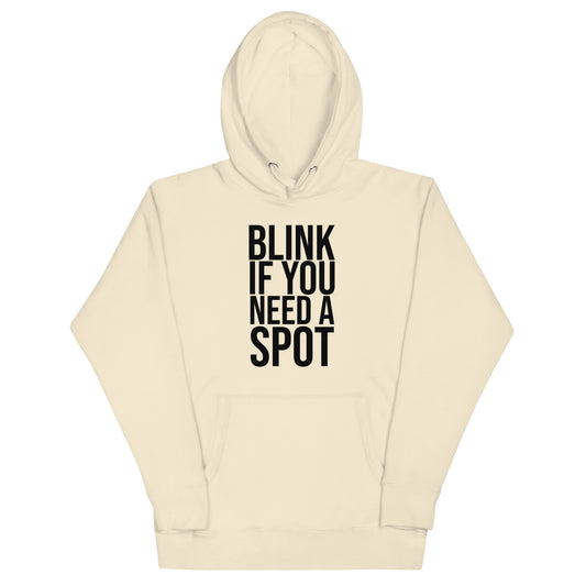 A man sits on a white cube wearing the Bougie Boiz Club Blink If You Need A Spot cream unisex hoodie, looking left with his left hand resting on the cube. Hes also wearing blue jeans.