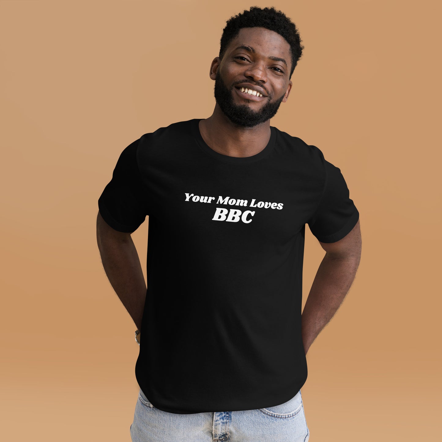 A smiling man combines humor and style in a navy Your Mom Loves BBC unisex T-shirt by Bougie Boiz Club, posing casually with hands in pockets against a neutral brown background. Ideal for gym or casual hangs, this look is both playful and relaxed.