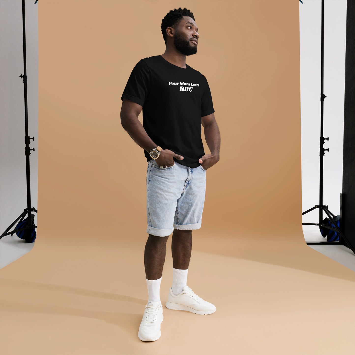 A smiling man combines humor and style in a navy Your Mom Loves BBC unisex T-shirt by Bougie Boiz Club, posing casually with hands in pockets against a neutral brown background. Ideal for gym or casual hangs, this look is both playful and relaxed.