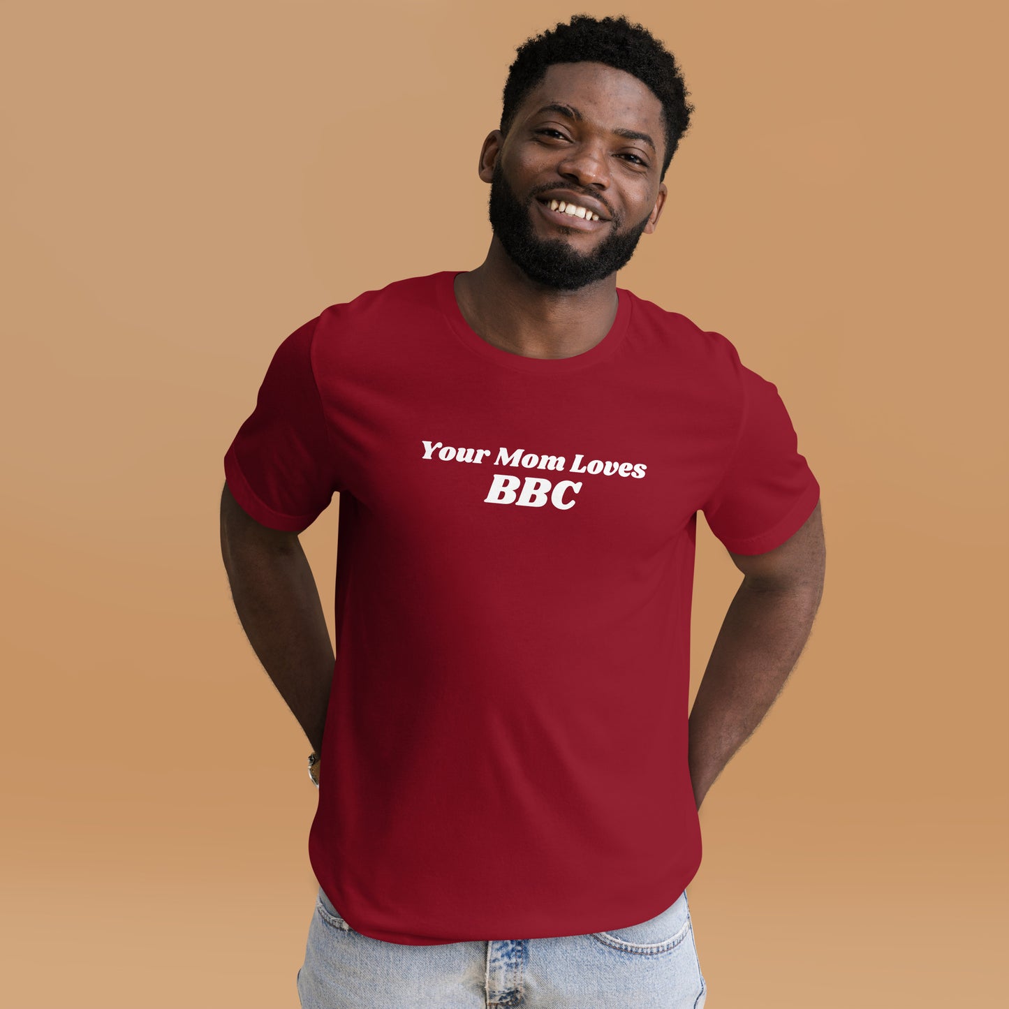 A smiling man combines humor and style in a navy Your Mom Loves BBC unisex T-shirt by Bougie Boiz Club, posing casually with hands in pockets against a neutral brown background. Ideal for gym or casual hangs, this look is both playful and relaxed.