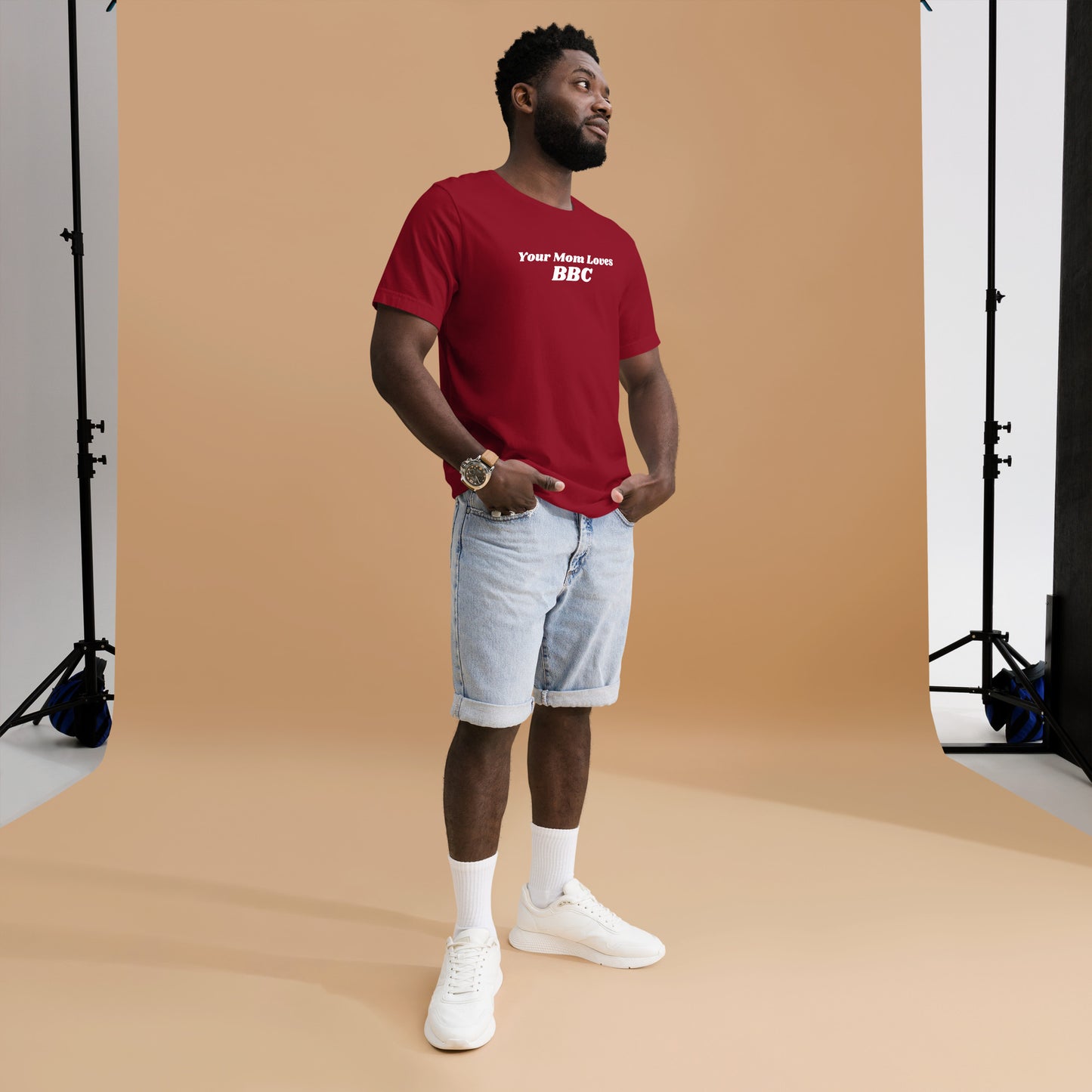 A smiling man combines humor and style in a navy Your Mom Loves BBC unisex T-shirt by Bougie Boiz Club, posing casually with hands in pockets against a neutral brown background. Ideal for gym or casual hangs, this look is both playful and relaxed.