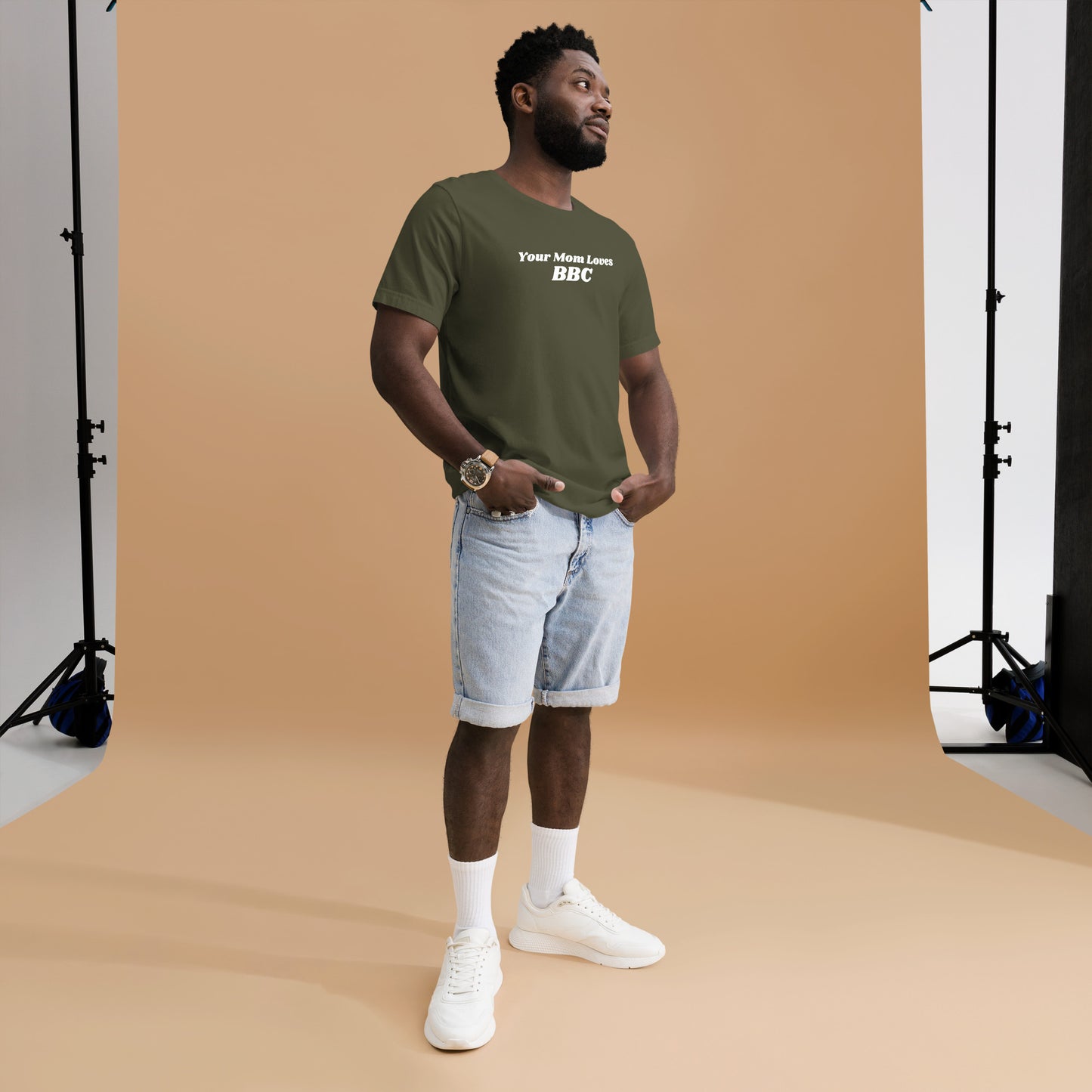 A smiling man combines humor and style in a navy Your Mom Loves BBC unisex T-shirt by Bougie Boiz Club, posing casually with hands in pockets against a neutral brown background. Ideal for gym or casual hangs, this look is both playful and relaxed.