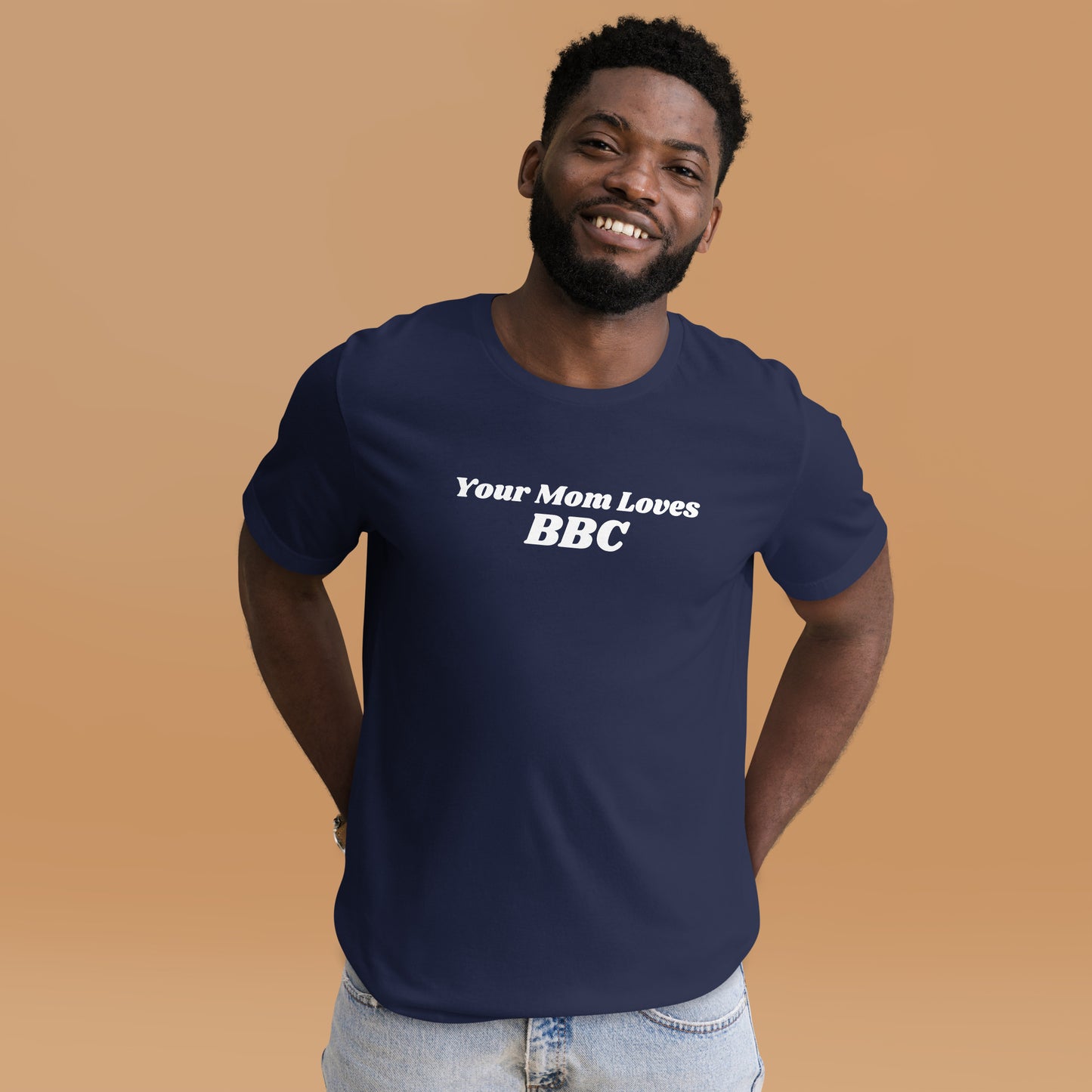 A smiling man combines humor and style in a navy Your Mom Loves BBC unisex T-shirt by Bougie Boiz Club, posing casually with hands in pockets against a neutral brown background. Ideal for gym or casual hangs, this look is both playful and relaxed.