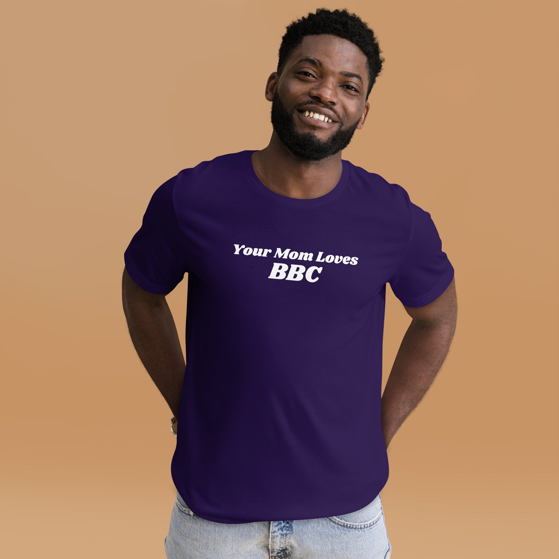 A smiling man combines humor and style in a navy Your Mom Loves BBC unisex T-shirt by Bougie Boiz Club, posing casually with hands in pockets against a neutral brown background. Ideal for gym or casual hangs, this look is both playful and relaxed.